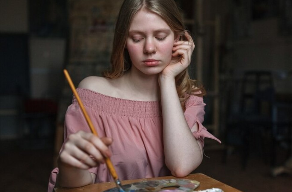 a sad girl in a pink dress is resting her head on her arm and mixing colors with a brush  on the palette