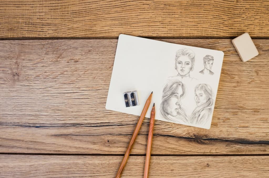 Open sketchbook with pencil portraits next to two wooden pencils and a sharpener on a wooden table