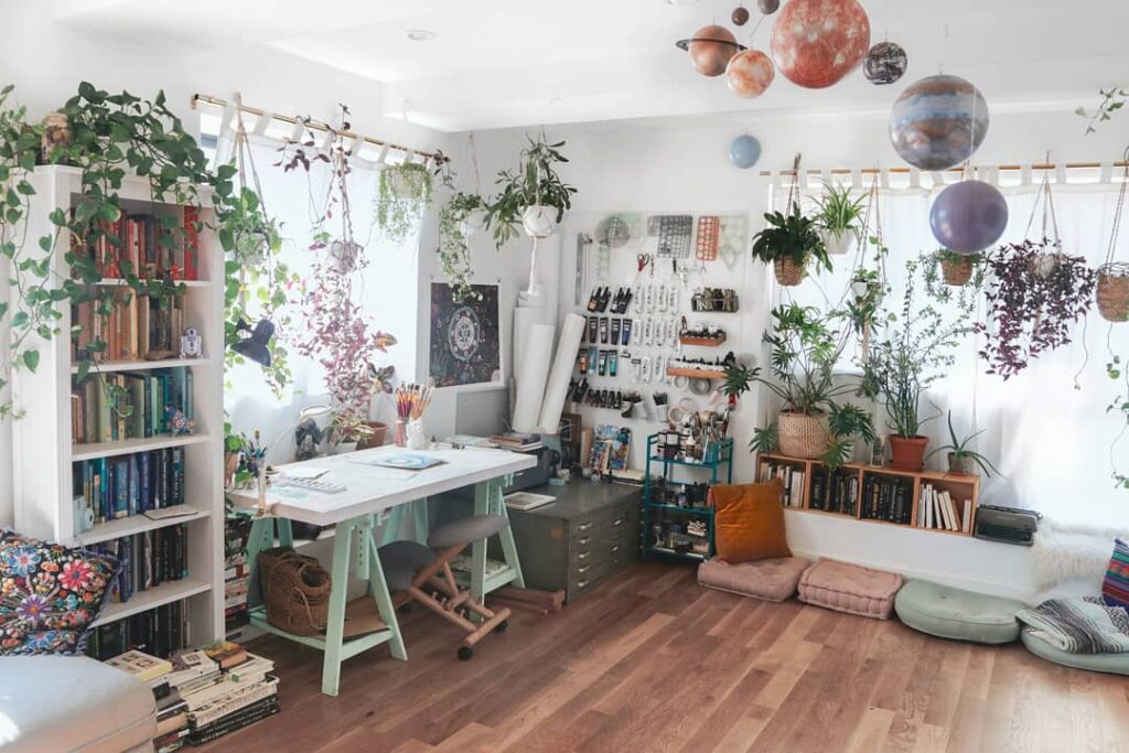 a small, bright room with lots of houseplants, a book rack, and an artist area