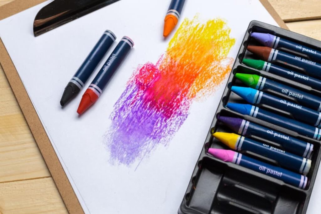 Oil pastels beside their colorful strokes on white paper, next to a clipboard