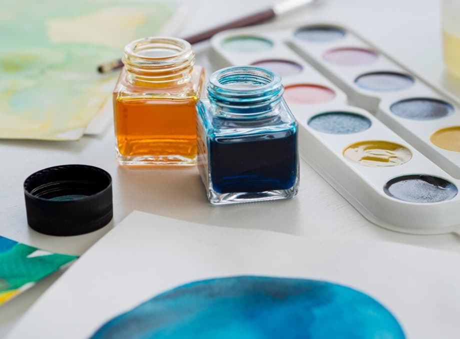 Open jars of orange and blue ink next to a watercolor palette on a white surface